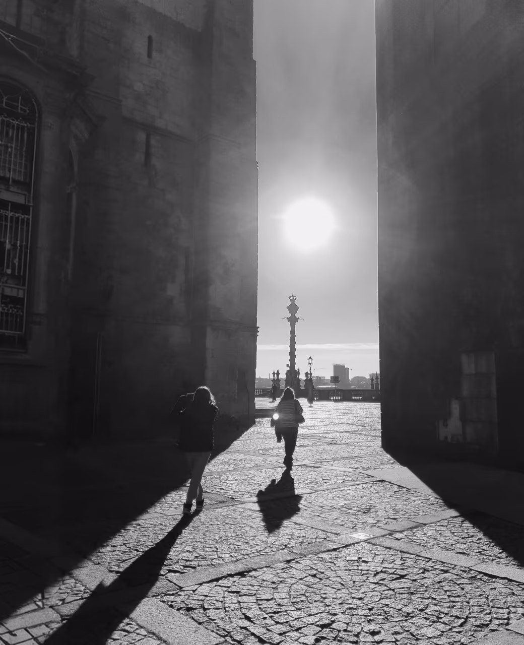 Two people walking through a sunlit cobblestone passage between tall buildings